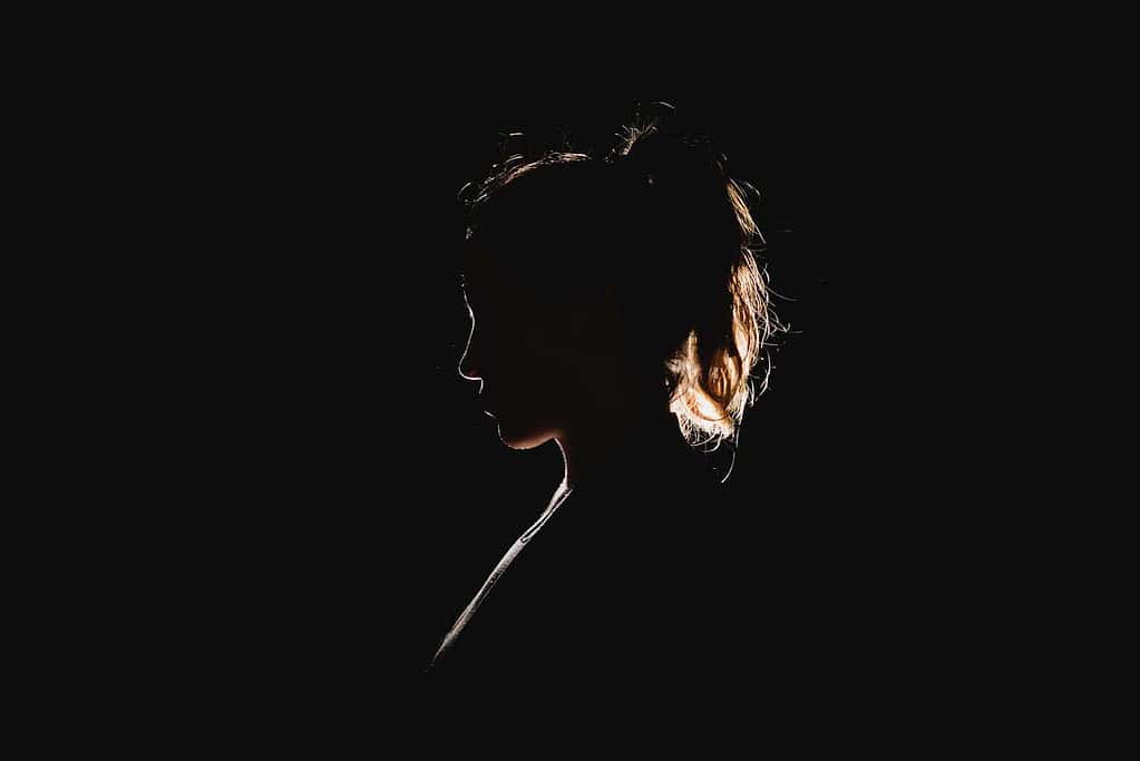 pitch black image with a woman standing and looking to the left and there is a spotlight shining behind her